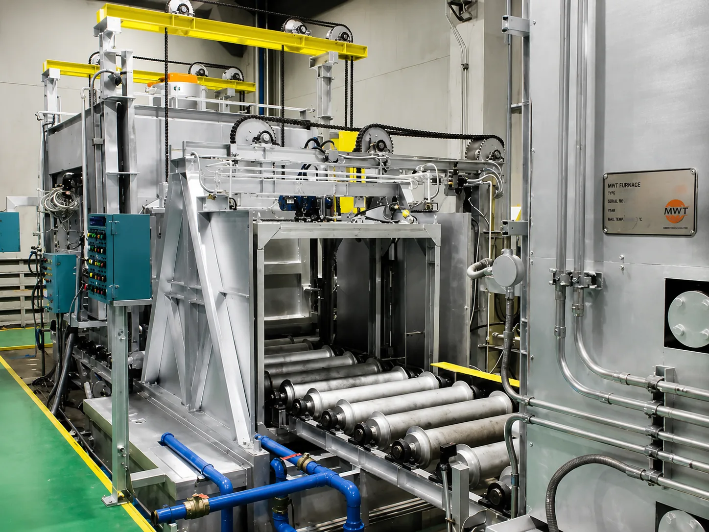 MWT T6 heat treatment furnace with roller conveyor entry, yellow lifting beams, chain drives, control panels and water piping inside a plant